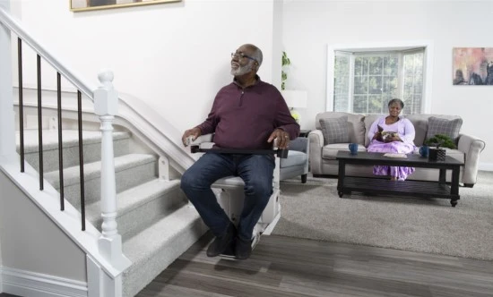 a man riding a curved stair lift with his wife sitting in the living room behind him watching