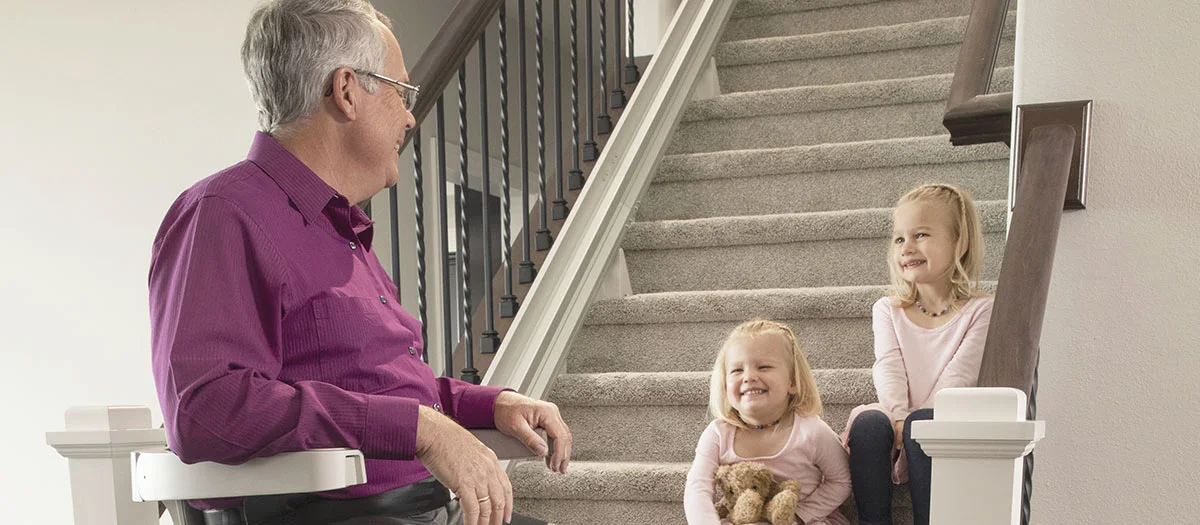 a man riding an indoor stair lift with kids sitting on the steps watching